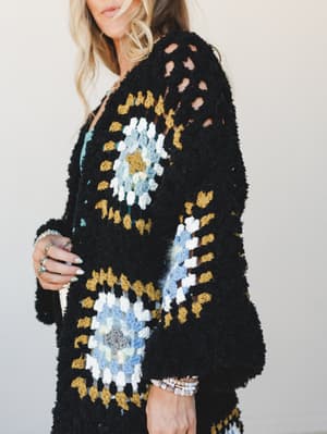 A close-up of a chunky black crochet cardigan with granny square panels in mustard, dusty blue, and white — worn over a light floral top and styled with stacked beaded bracelets and silver rings, giving it that perfectly lived-in, emotionally attache