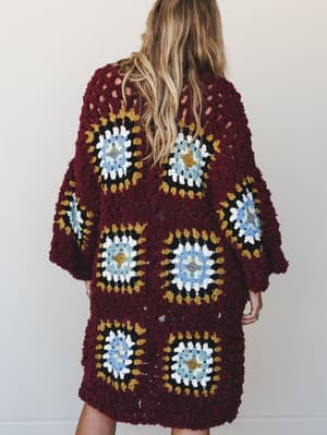 Shot from the back, a model with loose wavy hair wears a chunky longline granny square crochet cardigan in deep wine, the patchwork squares radiating dusty blue, mustard, black, and white — the kind of piece that looks like it took someone's grandmot