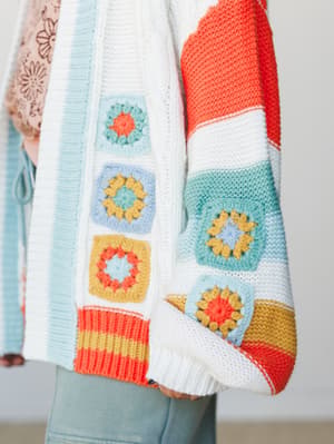 A close-up detail shot of a chunky knit cardigan in white with bold color-block stripes and hand-crocheted granny squares — the kind of piece that looks like it took someone's grandmother three winters to finish, in the best possible way.