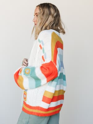 A cozy oversized cardigan in cream with bold retro-stripe colorblocking and granny square crochet patches — the kind of sweater that looks like it was made with love and worn everywhere.