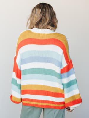 Shot from behind, a model wears an oversized rainbow-striped knit cardigan with wide bell sleeves — the kind of sweater that makes you want to drive somewhere with the windows down.