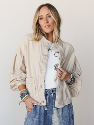 A relaxed, layered look built around a cream mixed-media bomber-style sweater jacket — knit ribbed placket meets woven body — worn open over a white graphic tiger tee and acid wash denim, with stacks of beaded and turquoise bracelets doing all the pe