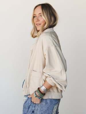 A relaxed side-profile shot of a model in a cream button-down bomber-style sweater layered over acid wash jeans, styled with a stack of boho beaded bracelets and a silver cuff — the kind of effortless outfit that looks like it came together without t