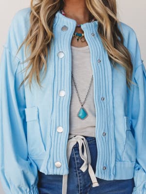 A soft sky blue button-down sweater jacket with ribbed knit detailing down the front is styled open over a knotted gray tee and dark denim, layered with turquoise statement jewelry — the kind of outfit that looks effortless but clearly wasn't an acci
