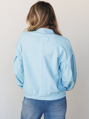 A back-view shot of a soft sky blue pullover sweatshirt with exposed seam detailing and a ribbed hem, paired with medium-wash jeans — easy, relaxed, and the kind of top you reach for without thinking.