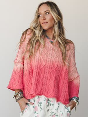 A breezy, lived-in cable-knit sweater in a coral-to-blush ombre styled with floral bottoms and stacked beaded bracelets — the kind of outfit that looks like it came together effortlessly but absolutely did not.