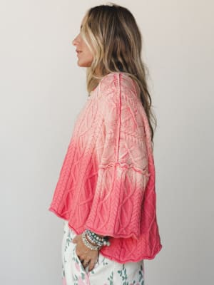 A side-profile shot of a model in a cable-knit sweater with a soft ombre wash from blush pink to coral, styled with floral pants and a stack of beaded bracelets — the kind of sweater you reach for when you want to feel put-together without trying too