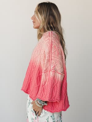 A side-profile shot of a model in a cable-knit sweater with a soft ombre wash from blush pink to coral, styled with floral pants and a stack of beaded bracelets — the kind of sweater you reach for when you want to feel put-together without trying too