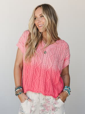 A bright coral cable-knit sweater top styled with floral wide-leg pants and stacked boho bracelets — the kind of outfit that looks like it was pulled together effortlessly but definitely wasn't.