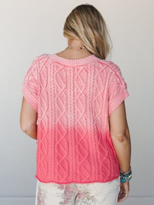 A back-view shot of a short-sleeve cable knit sweater in a soft pink-to-coral ombre wash, styled with floral pants and layered boho jewelry — the kind of top that makes a Saturday feel intentional.