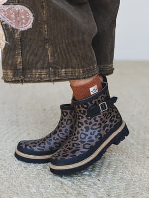 A close-up of leopard print rain boots with chunky lug soles and a side buckle strap, styled with rust ribbed socks and a dark corduroy midi skirt with floral patch appliqué — the kind of outfit that makes a rainy day feel like a personal choice.