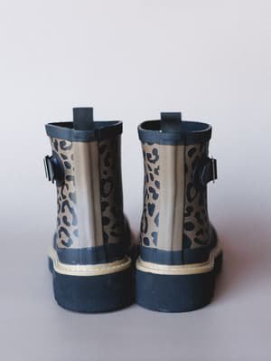 A pair of short leopard-print rain boots shot from the back, showing off their navy rubber sole and trim, side buckle hardware, and pull tab — practical with just enough personality to make a rainy day feel intentional.