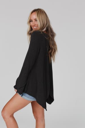 A woman with long, wavy blonde hair smiles over her shoulder, wearing the Golden Hour Long Bell Sleeve Top in Charcoal and denim shorts against a light gray background.