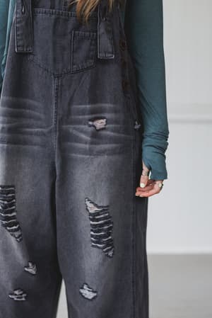 A person wears the High Hopes Slouchy Denim Overalls in black, featuring distressed details and an oversize silhouette from torso to upper legs, layered over a teal long-sleeved shirt indoors.
