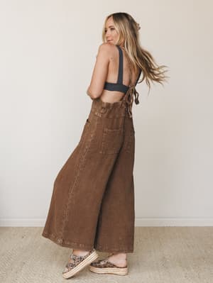 Shot from behind with a glance-back moment, this model wears wide-leg chocolate brown overalls in a textured woven fabric, styled open-back with tie straps over a dark sports bra — easy, earthy, and a little unexpected.
