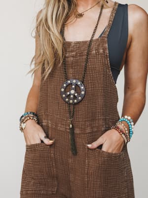 A close-up torso shot of a model in chocolate brown gauze overalls layered over a black tank, styled with a statement peace sign pendant necklace and stacked beaded bracelets — effortlessly lived-in with a free-spirited edge.