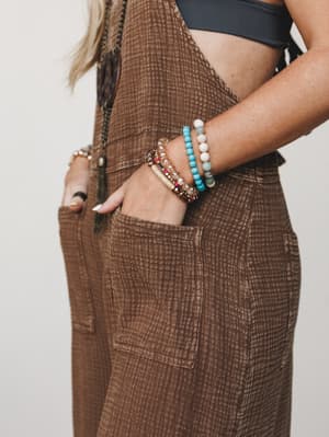 A close-up detail shot highlighting a stacked beaded bracelet set against the textured chocolate brown overalls — hand tucked in a front patch pocket, layered jewelry doing all the talking.