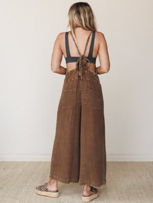 A model shot from behind showing wide-leg overalls in a warm chocolate brown textured fabric, styled open-back with a black bralette and espadrille platform sandals — effortlessly relaxed with just enough personality.