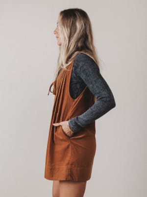 Woman wearing a brown pinafore dress over a dark long-sleeve shirt against a plain background