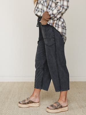 A cropped side-angle shot showing acid-washed black barrel pants styled with a frayed plaid top, a layered waffle knit sleeve, and leopard print platform sandals — relaxed and a little lived-in, in the best way.