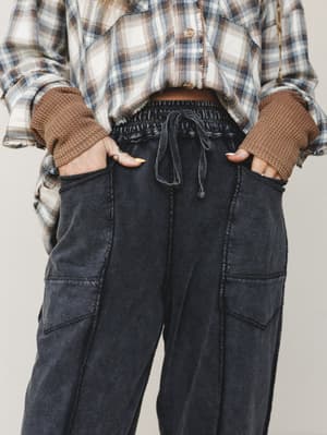 A close-up waist-down shot of washed black barrel-leg cargo pants styled with a layered plaid flannel and waffle knit — the kind of effortless, thrown-together outfit that somehow looks completely intentional.