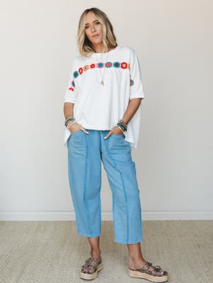 An oversized white tee with a colorful crochet floral trim is paired with relaxed barrel-leg pants in a soft sky blue — the kind of effortless, feel-good outfit that makes a Tuesday errand run look intentional.