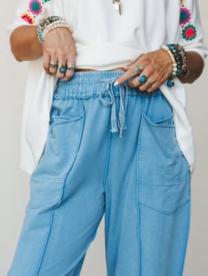 A close-up mid-shot centered on soft blue barrel-leg pants with an elasticized drawstring waist, styled with a white embroidered top and layered silver and turquoise jewelry — relaxed and expressive from the waist down.