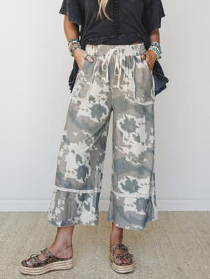 Wide-leg cropped pants in a soft, faded camo print styled with a washed black tee, stacked turquoise bracelets, and leopard platform sandals — relaxed and a little unexpected in the best way.