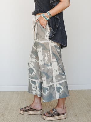 A side-profile shot of wide-leg camo-print crop pants in faded olive and cream, styled with a washed black ruffle top, leopard print platform sandals, and a stack of turquoise beaded bracelets — the kind of effortless, layered look that feels persona