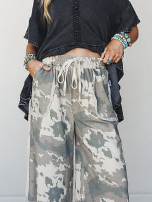 A mid-shot of wide-leg drawstring pants in a soft camo-style tie-dye print styled with a washed black crop tee, denim jacket draped over the arm, and stacked turquoise and beaded jewelry — relaxed, expressive, and very much a 'Saturday with nowhere t