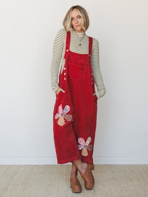 A wide-leg corduroy overall in bold red worn over a sage striped turtleneck, with patchwork floral appliqués on the legs and clogs — the kind of outfit that feels like a thrifted treasure and a whole personality at once.