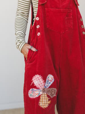 A close-up of red corduroy overalls layered over a sage stripe long-sleeve top, with a charming patchwork floral appliqué on the leg — the kind of piece that feels like it came from someone's very cool grandmother's closet.
