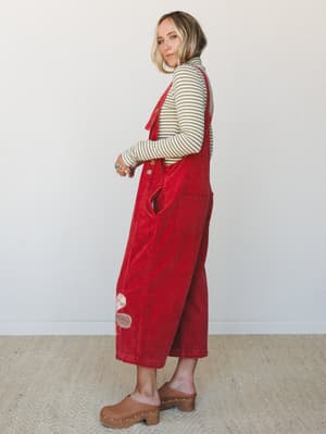A model styled in red corduroy wide-leg overalls layered over an olive striped long-sleeve turtleneck, shot in profile against a clean white studio wall — the kind of outfit that feels vintage-inspired and totally lived-in at the same time.
