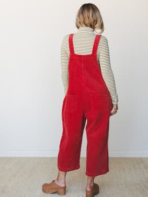 A back-facing model wears wide-leg red corduroy overalls over a striped turtleneck, styled with tan clogs — the kind of outfit that feels effortless and a little nostalgic all at once.