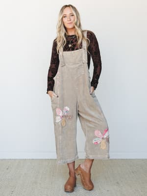A wide-leg corduroy overall in washed latte worn over a dark floral long-sleeve top, styled with wooden clogs and layered necklaces — the kind of outfit that looks like it came together effortlessly but absolutely did not.