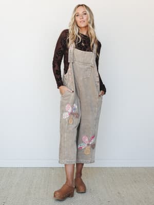 Corduroy overalls in a warm latte tone, dressed up with a dark floral velvet long-sleeve and wood-platform clogs — the patchwork flower appliqués make it feel like something your coolest aunt would have saved from 1974.