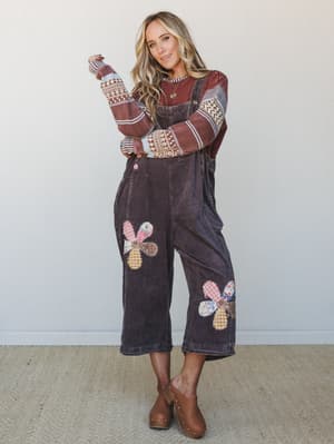 A patchwork-sleeved layered knit tops washed corduroy overalls with charming fabric flower appliqués — the kind of outfit that looks like it was styled by someone who genuinely loves getting dressed.