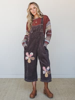 A wide-leg corduroy overall in deep mocha is layered over a patchwork-print long sleeve top, finished with wooden clogs and patchwork flower appliqués on each leg — the kind of outfit that feels like it came from a really good thrift run and a craft