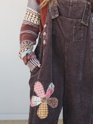 A close-up torso shot of corduroy overalls in deep mocha, layered over a patterned fair isle sweater — the standout detail is a patchwork flower appliqué made from mismatched plaid and floral fabric scraps, the kind of thing that looks like it came f