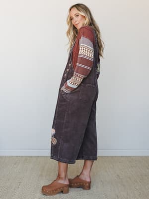 A side-profile shot of a model in wide-leg corduroy overalls layered over a fair isle mixed-stripe sweater, styled with wood-platform clogs — the whole look has that effortless, lived-in autumn energy.