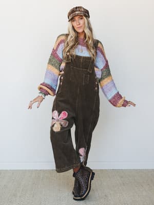A full-body model shot of a layered boho outfit built around olive corduroy overalls with patchwork flower appliqués, worn over a multicolor stripe knit sweater and finished with a plaid newsboy cap and leopard print boots — the kind of look that fee