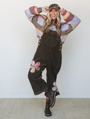 A smiling model layers a multicolor striped knit sweater under wide-leg corduroy overalls with patchwork flower appliqués, topped with a plaid newsboy cap and leopard print boots — the kind of outfit that looks like it was assembled with joy and a ve