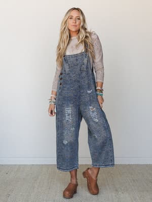 A relaxed, full-body shot of a model wearing wide-leg leopard-print denim overalls over a sheer lace long-sleeve top, styled with stacked beaded bracelets and brown studded clogs — the kind of outfit that feels effortlessly put-together without tryin