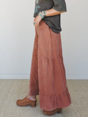 A side-profile shot of wide-leg tiered pants in a washed brick tone, styled with a faded graphic tee, stacked turquoise bracelets, and wood clogs — the kind of outfit that looks effortless but is clearly thought through.