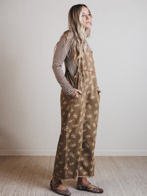 Woman wearing a brown floral jumpsuit with a striped shirt underneath, standing against a plain wall.