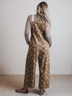 Person wearing a brown floral overalls with a striped shirt underneath, standing against a white wall.