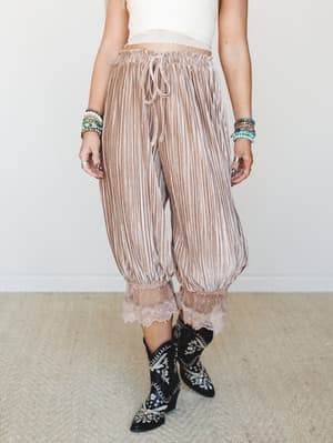 Flowy pleated trousers in warm mocha tones with a lace hem finish, styled with a cropped lace-trim top, embroidered black cowboy boots, and stacked beaded bracelets — the kind of outfit that looks like it came together effortlessly but definitely did