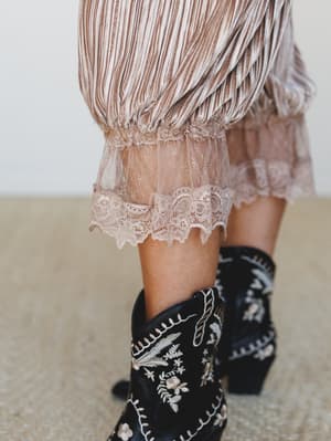 A close-up detail shot of a pleated mocha trouser with a delicate lace hem paired with black embroidered cowboy boots — the kind of contrast that feels intentional and a little magic.