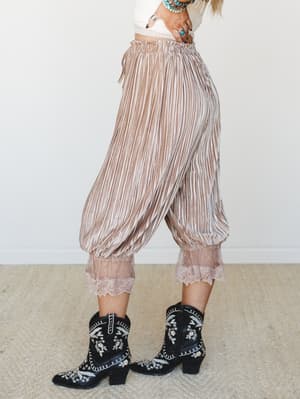Pleated velvet-finish trousers in a warm mocha stripe with a banded lace hem, styled with a cropped cream top, embroidered black cowboy boots, and turquoise stacked bracelets — the kind of outfit that makes a statement without trying too hard.