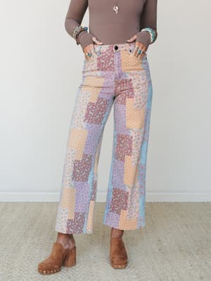 A cropped model shot showing wide-leg patchwork denim pants in a dreamy mix of pastel floral and dotted prints, styled with a taupe long-sleeve bodysuit, stacked boho bracelets, and suede platform clogs — equal parts vintage quilt energy and effortle